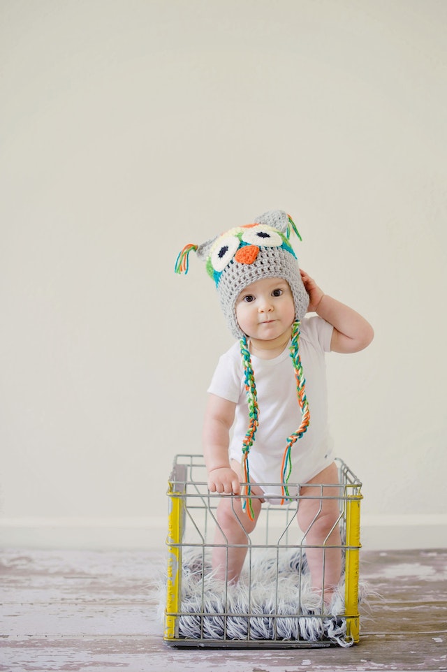 Kleinkind mit Mütze steht in einem Drahtkorb mit Fell wie in einem Laufgitter